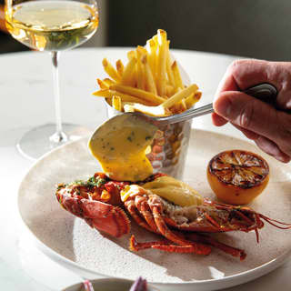 A guest spoons herby Hollondaise onto grilled lobster served with seared lemon and frites, with side salad and white wine.