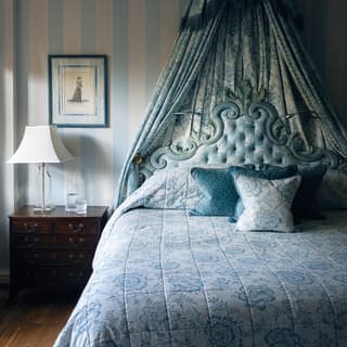 Pale blue and white vertical candy striped wallpaper fills the walls behind a king bed with ornate padded pale blue bedhead
