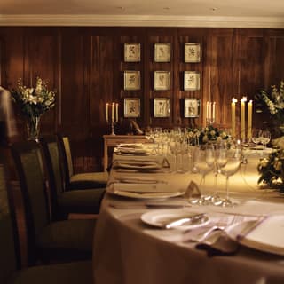 Nine framed botanical drawings hang from a wood panelled wall. An elegant table is set for dinner, decorated with white roses
