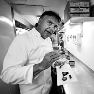 Raymond Blanc uses his hand to emphasise a point as he speaks to someone off camera about a dish of food he is holding